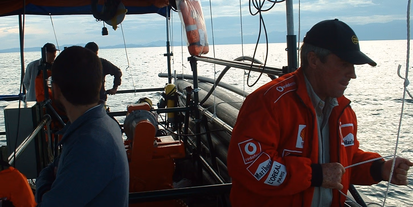 A man in a red jacket and black cap holds a rope on a boat on the water, with other people working in the background.