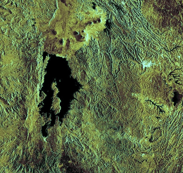 Satellite image showing rugged green terrain with a large, dark, elongated lake surrounded by mountainous ridges and valleys.
