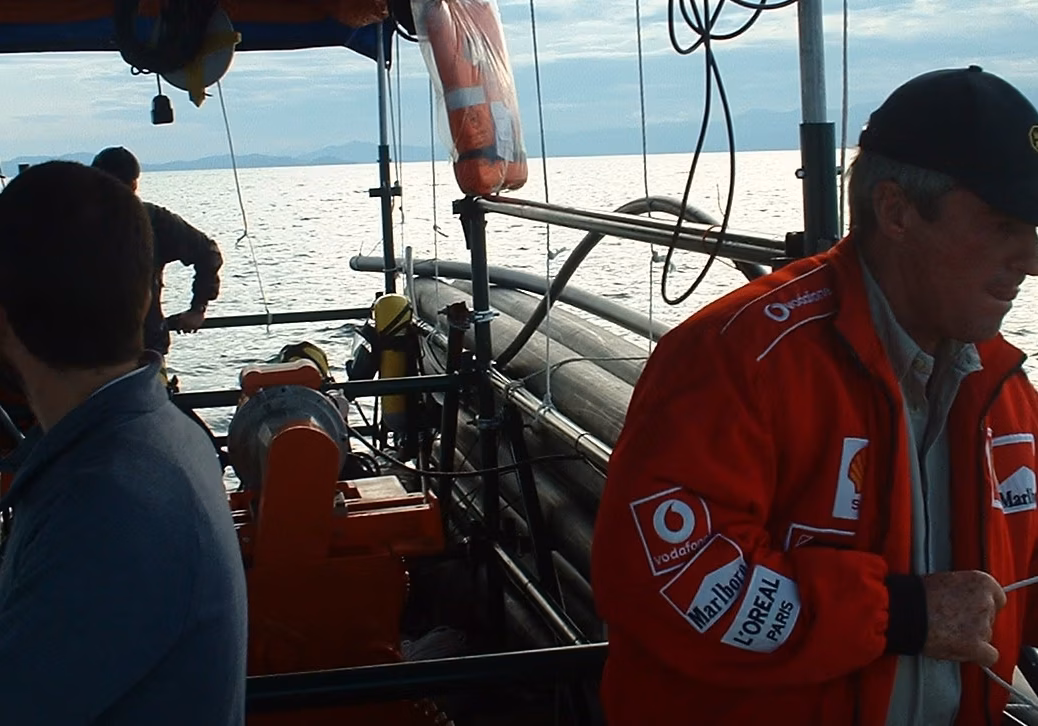 A man in a red jacket and black cap holds a rope on a boat on the water, with other people working in the background.